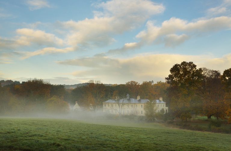 A large white house sits among autumn trees, partially shrouded in morning mist, with a green grassy field in the foreground and a blue sky with scattered clouds above.