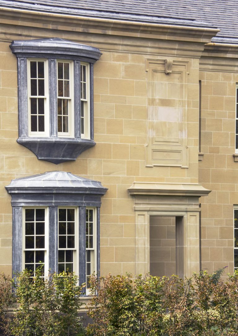 A tan stone building with decorative trim features two large windows with blue-gray frames and a third space that appears bricked over, creating a “fake” window above a door-like opening. Green shrubs grow along the bottom of the image.