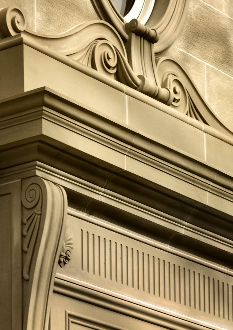 A close-up of an ornate beige stone building facade features detailed classical architectural elements, including scrollwork, fluted moldings, and decorative carvings, with soft lighting highlighting the intricate textures and craftsmanship.