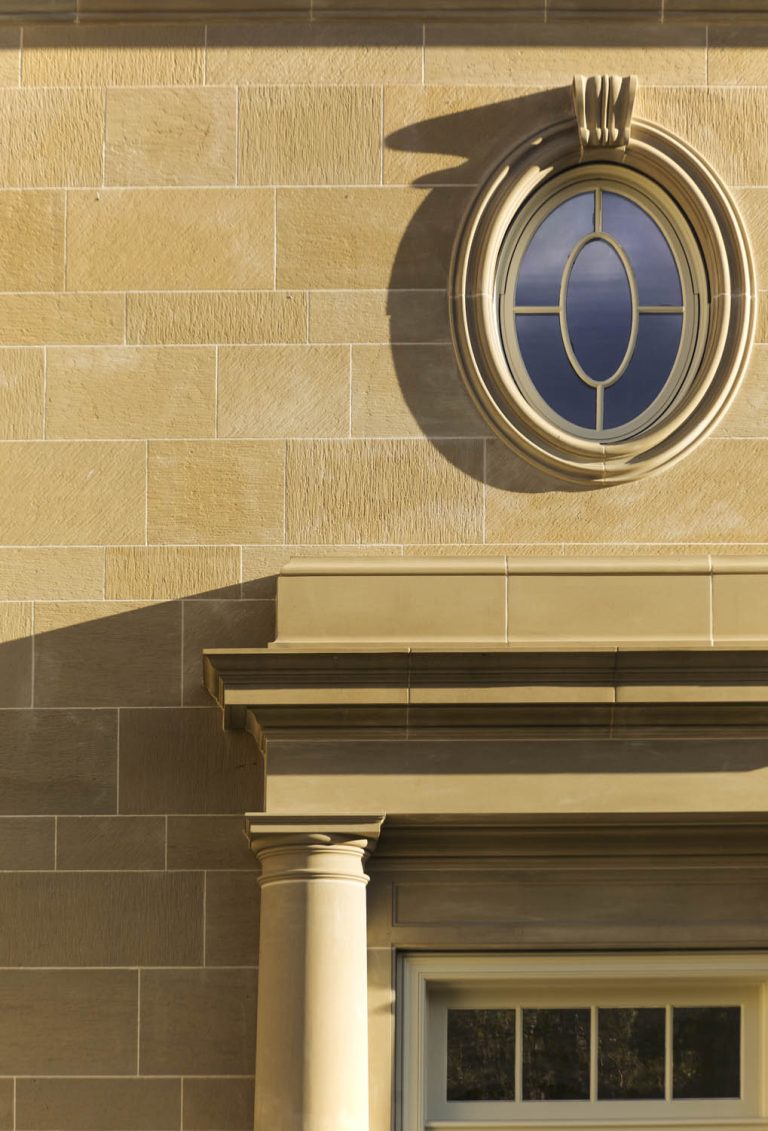 A beige stone building facade with rectangular blocks, an ornate round window with a cross design and deep blue glass, and part of a columned doorway topped by a detailed cornice, all illuminated by sunlight.
