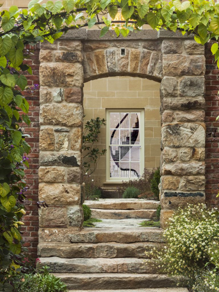 A stone archway with steps leads to a window framed by beige bricks. The window has a black and white geometric design. Green vines and leafy plants grow around the arch and pathway, adding a lush, inviting touch to the garden entrance.