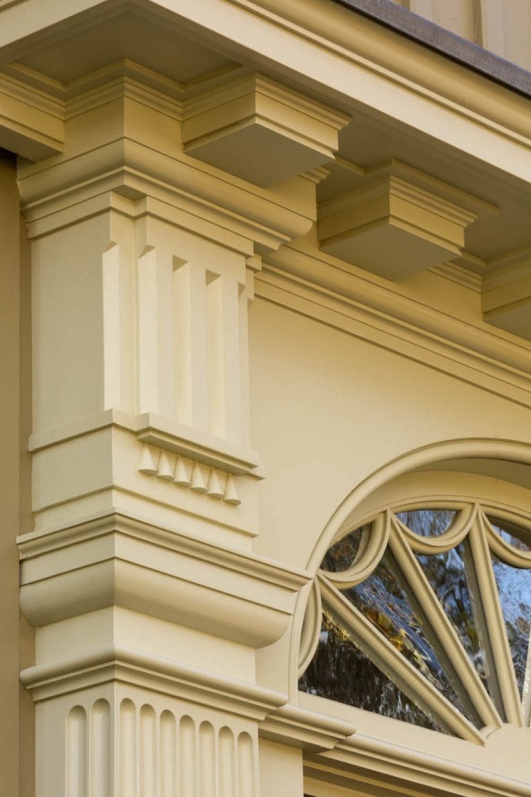Close-up of ornate, classical architectural molding on a yellow building. Design features rectangular columns, layered cornices, geometric details, and an arched window with radiating spokes reflecting trees and sky.