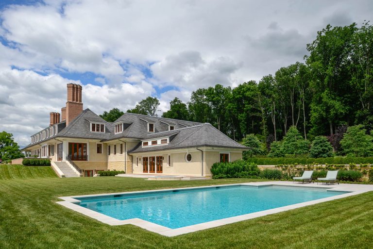 A large, light yellow house with gray shingles and multiple chimneys sits beside a rectangular swimming pool. The backyard features manicured grass, poolside lounge chairs, and is bordered by lush green trees under a partly cloudy sky.
