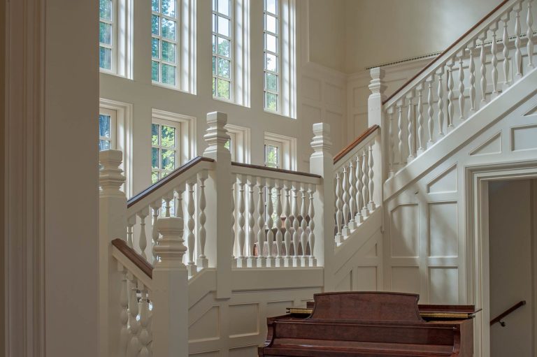 A sunlit staircase with white paneling and elegant spindles rises beside tall multipaned windows. Sunlight streams through the glass, casting shadows on the walls. At the bottom, a polished wooden piano sits in the foreground.