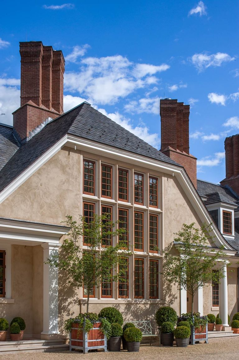 A large house with cream-colored walls, tall red brick chimneys, and many rectangular windows framed in wood. Potted trees and shrubs line the front entry. The sky is blue with scattered white clouds, and sunlight brightens the scene.