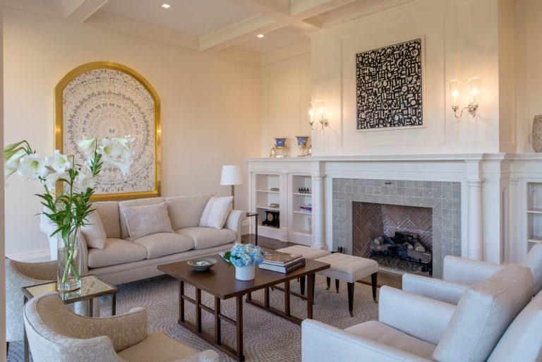Elegant living room with light-colored sofas and chairs arranged around a dark wood coffee table. A fireplace with herringbone tile, built-in shelves, and art above is the focal point. Floral arrangements and soft lighting enhance the cozy, sophisticated atmosphere.
