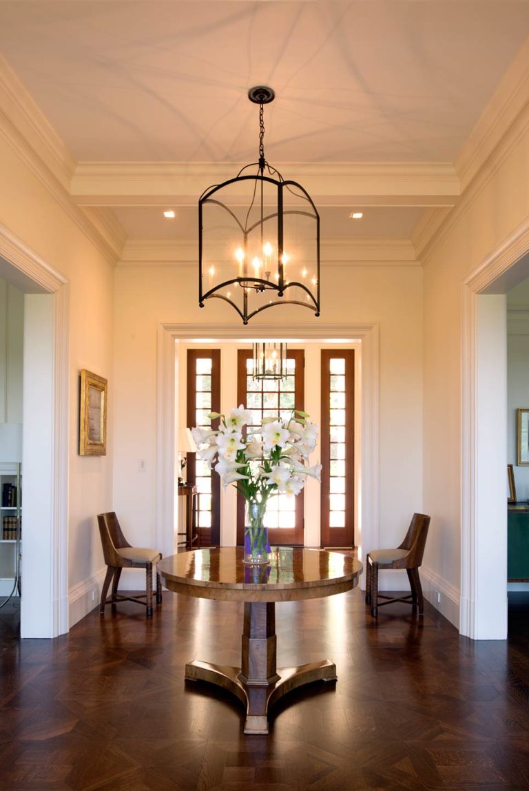 Elegant entryway with dark wood floors, a round wood table with a vase of white flowers at the center, two wooden chairs against cream walls, and a large glass pendant light overhead. Double wooden doors with glass panels let in natural light.