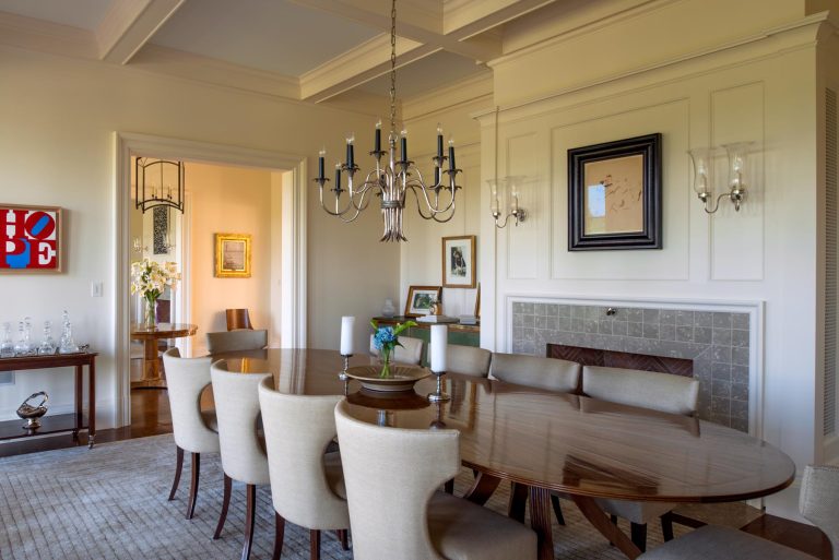Elegant dining room with a long polished wooden table surrounded by upholstered chairs. A classic chandelier hangs above. The room features wall art, a decorative fireplace, and soft natural lighting. Neutral tones and neatly arranged decor create a refined ambiance.