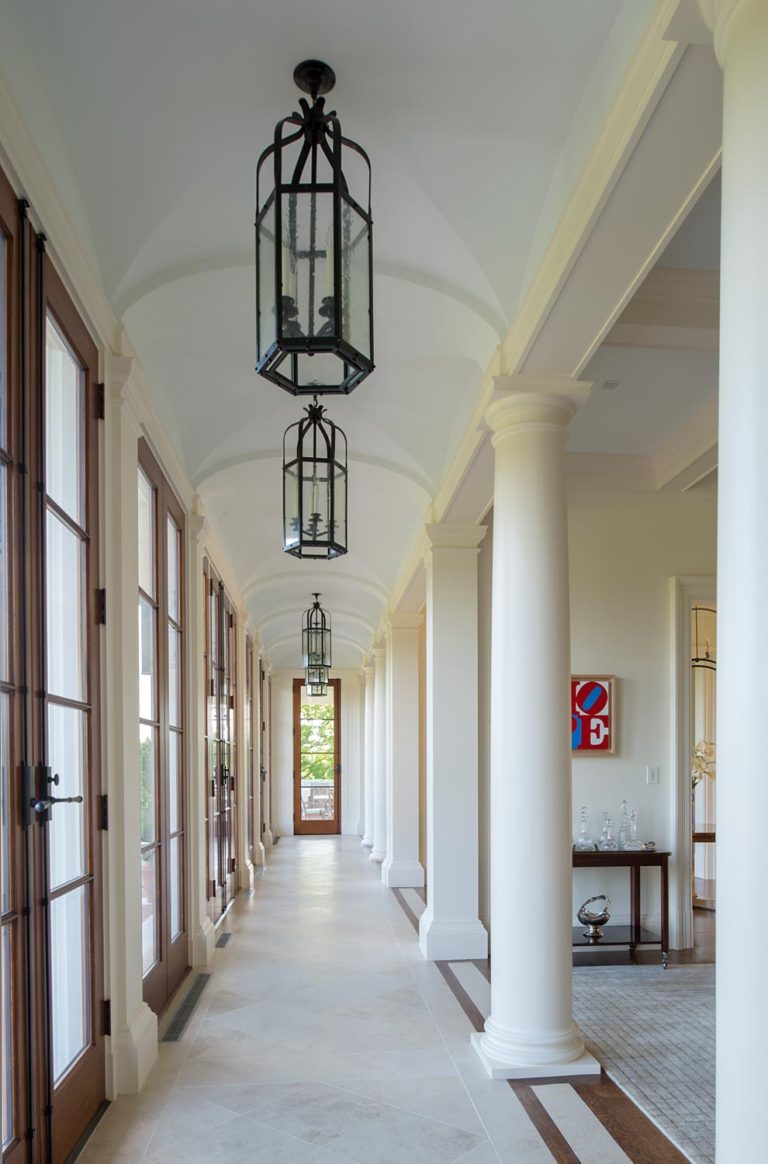 A bright hallway with high arched ceilings, tall white columns, and hanging black lantern lights. Large wooden-framed glass doors line the left side, allowing sunlight in. A small table and abstract art are visible at the end of the hallway on the right.