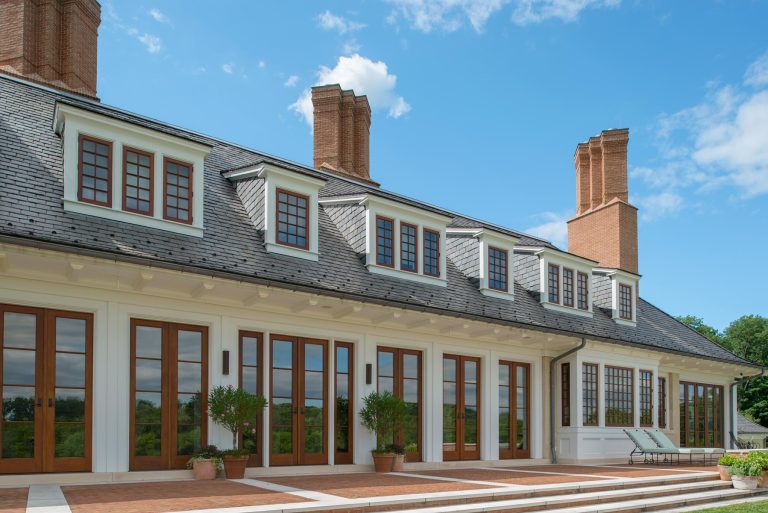 Large, elegant house with multiple tall brick chimneys, dark slate roof, many wooden-framed windows and doors, and a spacious patio with potted plants and lounge chairs. Bright blue sky with scattered clouds in the background.