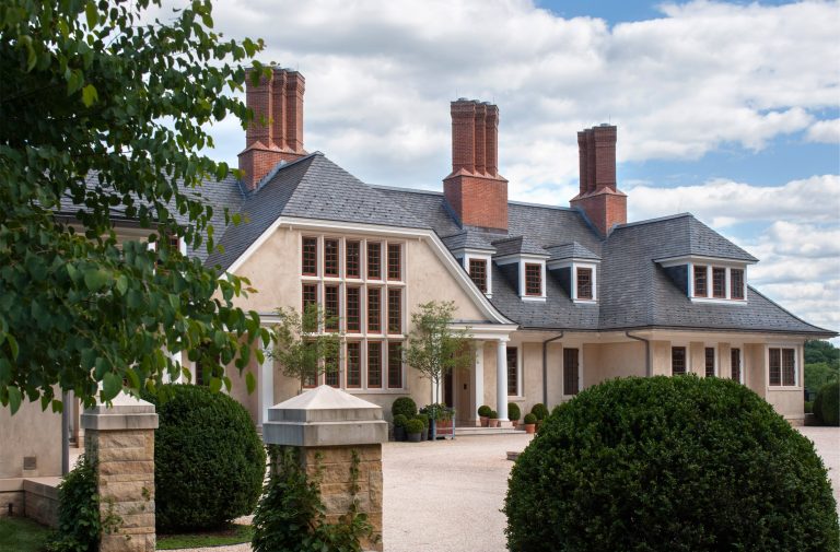 Large beige mansion with multiple tall red brick chimneys, slate roof, and many windows. Circular driveway bordered by manicured bushes and stone pillars. Trees and a partly cloudy sky are in the background.