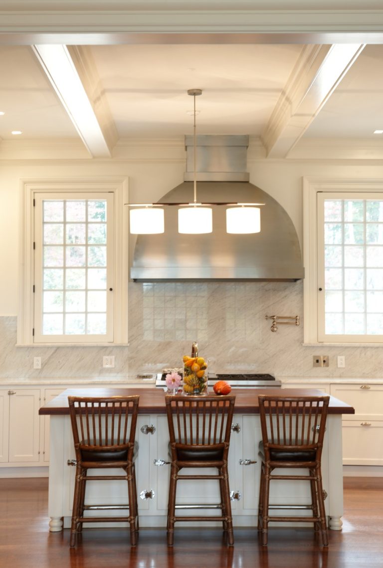 A modern kitchen with a center island, three wooden barstools, a fruit bowl, white cabinetry, stainless steel stove hood, and two large windows. A rectangular light fixture hangs above the island, and the floor is dark wood.