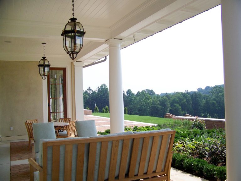 A covered patio with white columns and hanging lanterns overlooks a lush garden and distant trees. Wooden outdoor furniture with light blue cushions sits on a brick floor under the roof, creating a relaxing seating area.
