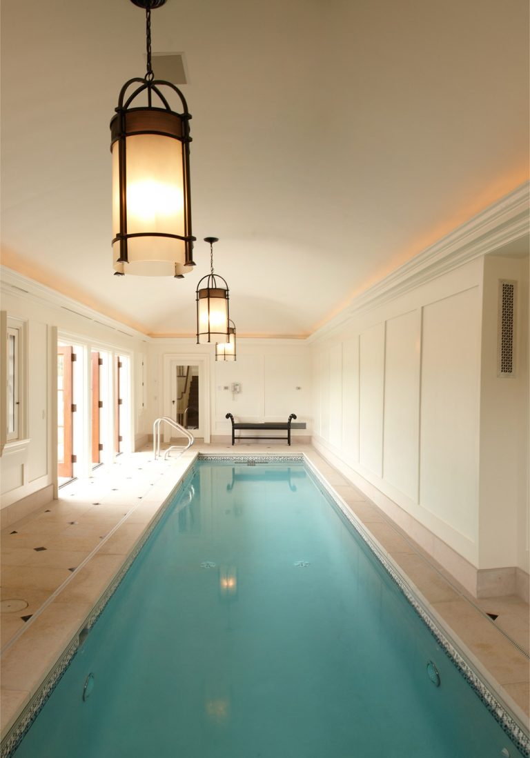 A narrow indoor swimming pool with calm blue water stretches along a cream-colored room. Large hanging lantern lights illuminate the space. Windows and glass doors line the left side, and the ceiling is vaulted. The design is elegant and tranquil.