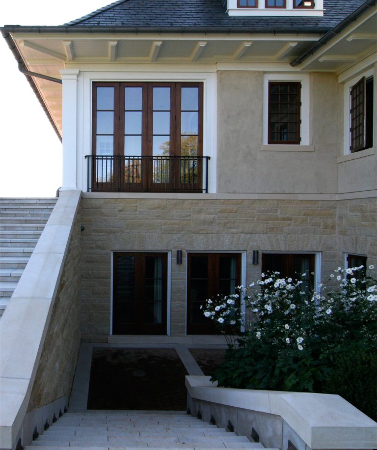 A stone stairway with white railings leads to a beige house with large wooden-framed windows and doors. White flowers and green plants border the lower entrance. The upper level features a small balcony with a black railing.