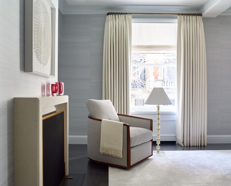 A modern, minimal living room features a light gray armchair with a beige throw, beside a white fireplace and a glass lamp. Cream curtains frame a large window, illuminating the neutral-toned space with soft natural light. A textured artwork hangs above the mantel.