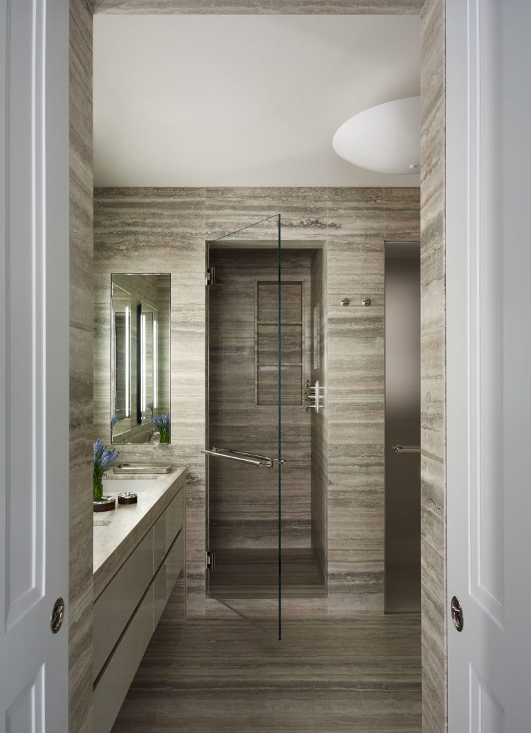 Modern bathroom with beige stone walls and floors, a glass shower door, a built-in shower shelf, large mirror, sleek vanity with drawers, white countertop, and small decorative items including a vase with blue flowers. White double doors frame the entry.