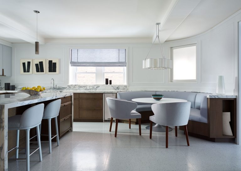 A modern kitchen features marble countertops, a breakfast bar with light blue stools, a built-in banquette with a round table and gray chairs, wood cabinetry, and neutral decor. Large windows bring in natural light, highlighting the clean, minimalist design.