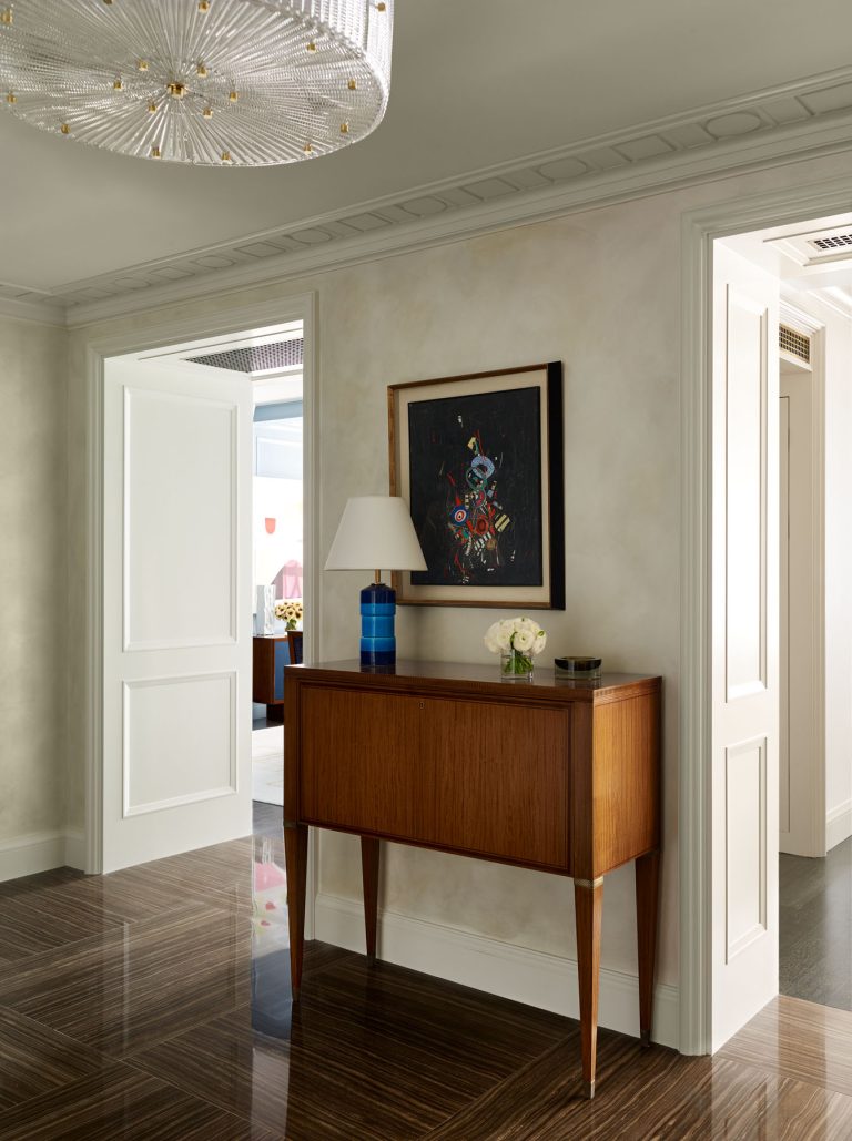 A modern hallway features glossy dark brown floors, soft beige walls, and a wood console with tapered legs. On the console sit a blue and white lamp, white flowers, and a dish. Above hangs a colorful abstract painting. A chandelier is on the ceiling.