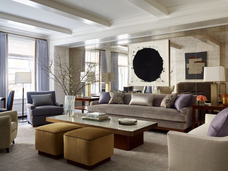 Elegant living room with a gray sofa, armchair, two tan ottomans, and a glass coffee table. Large mirror behind the sofa reflects light. Modern black-and-white artwork hangs above the sofa. Tall vase with branches sits on the coffee table. Neutral tones throughout.