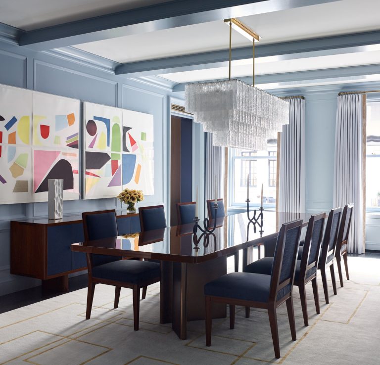A modern dining room with light blue walls, a dark wooden table, eight dark blue upholstered chairs, and a large rectangular crystal chandelier. Abstract colorful artwork hangs on the wall, and a sideboard with a vase and flowers sits to the left.