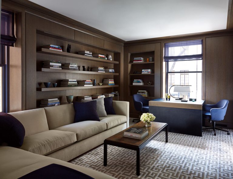 A modern home office features a beige sofa with blue and gray cushions, a dark coffee table with flowers, built-in wooden bookshelves filled with books and decor, a geometric-patterned rug, a sleek desk by two windows, and blue upholstered chairs. Natural light fills the room.