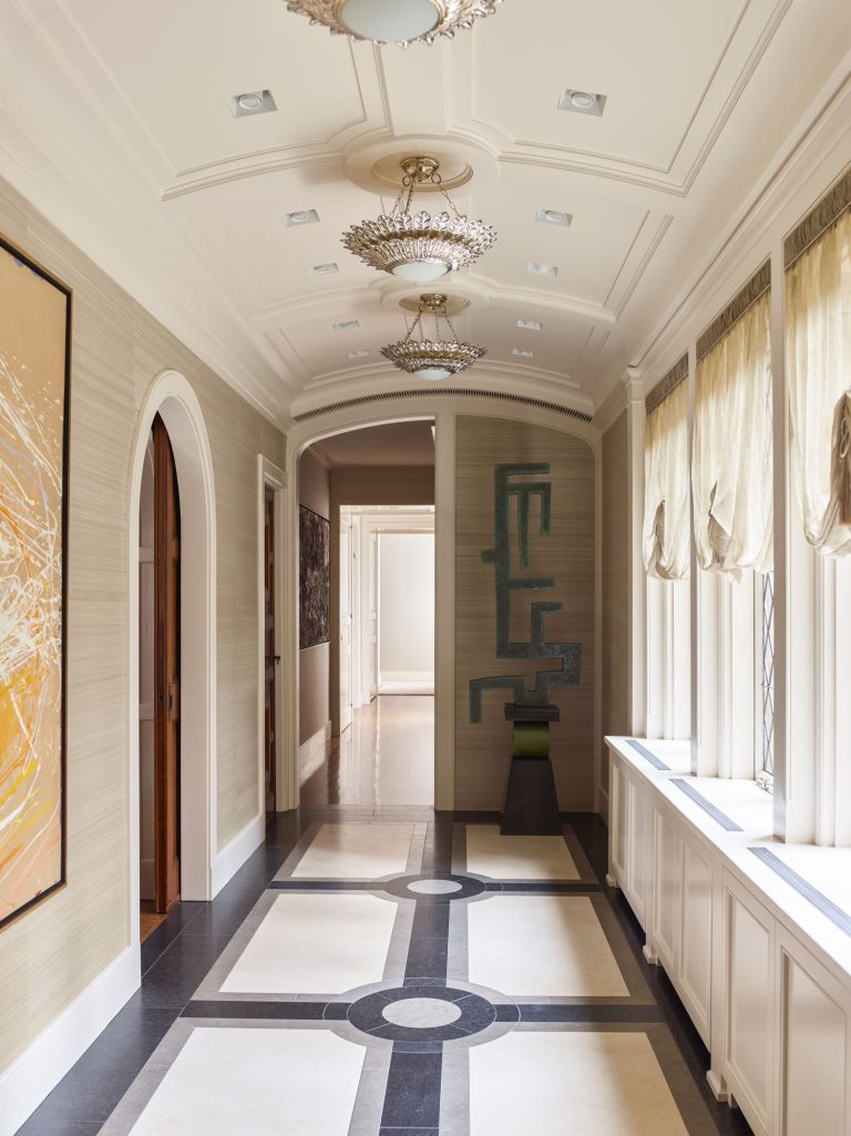 A bright hallway with cream walls, tray ceiling, ornate chandeliers, and large windows with beige Roman shades. The floor has a geometric tile pattern, and abstract art hangs on the walls. A green abstract sculpture stands at the end of the hall.
