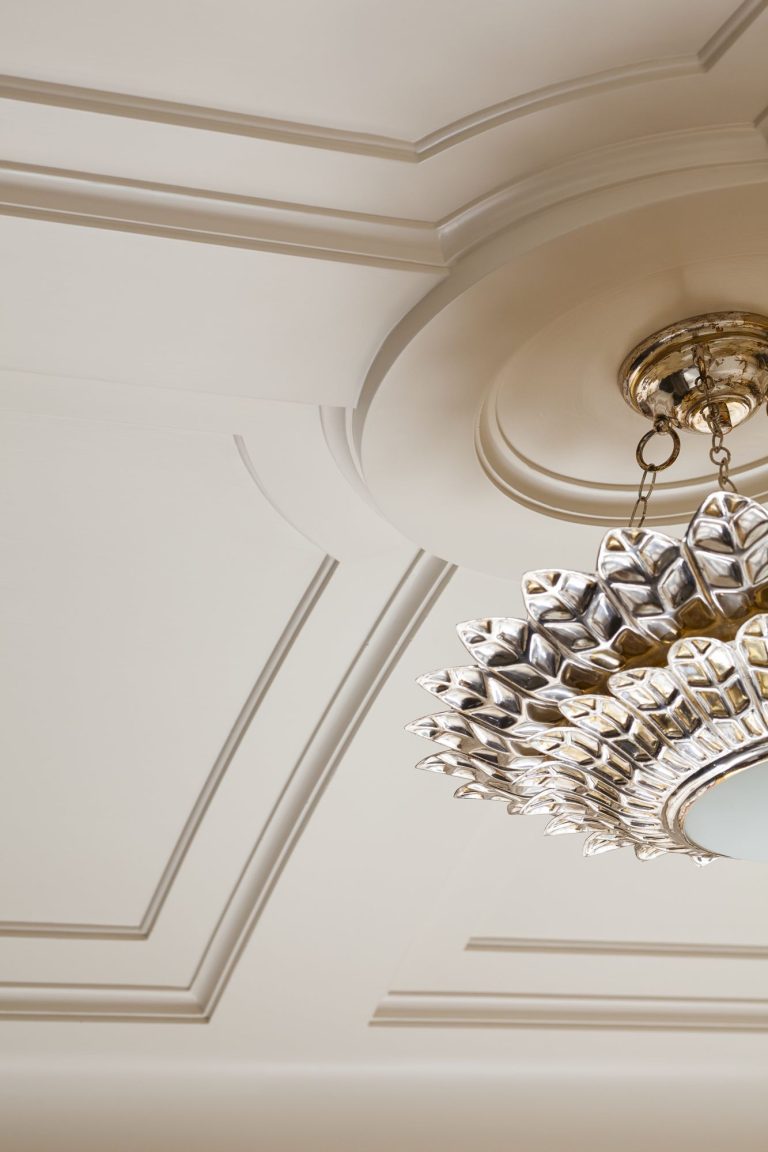 A close-up of an ornate ceiling with elegant moulding details and a decorative chandelier featuring metallic, leaf-shaped elements, hanging from a round ceiling medallion. The overall color palette is cream and gold.