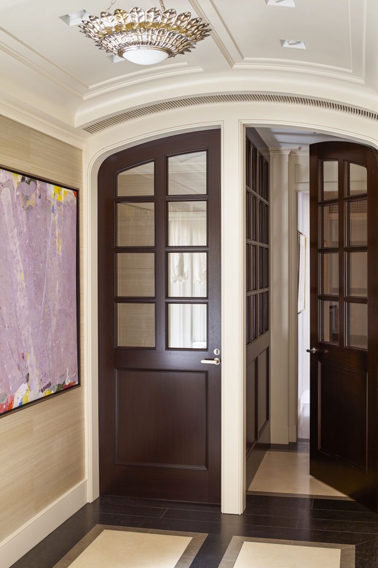A hallway with a large, dark wood door with glass panels, slightly open to reveal another matching door. The walls are beige, one featuring an abstract painting with pink and purple hues. The ceiling has an ornate light fixture. The floor is checkered beige and dark brown.