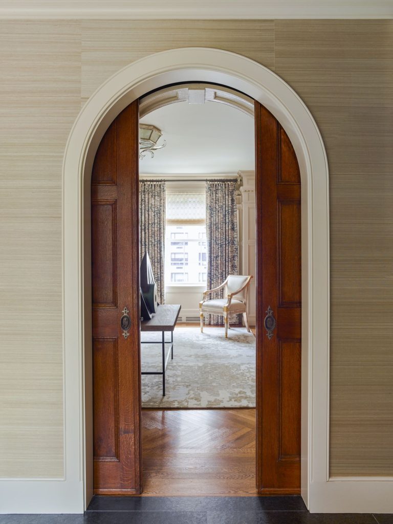A wooden arched doorway with sliding doors opens into a bright living room with a large window, patterned curtains, a beige rug, a sofa, and a wooden chair; sunlight streams in, highlighting hardwood floors and neutral-toned walls.