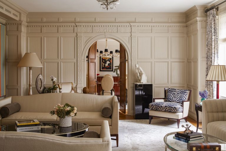 Elegant living room with beige paneled walls, cream sofas, glass coffee tables, books, and a zebra-print chair. An arched doorway leads to a dining room with a round table, chairs, a modern chandelier, artwork, and a white sculpture on a black pedestal.