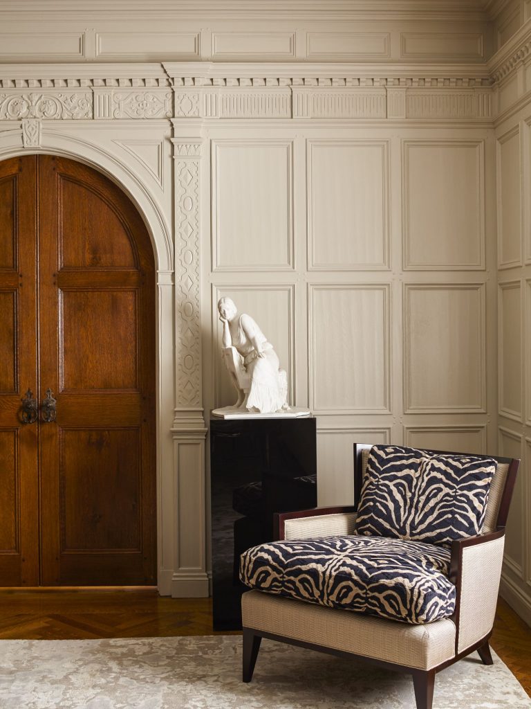 A beige-paneled room features a carved wooden double door, a white marble sculpture of a seated woman on a black pedestal, and an armchair with dark wooden arms and zebra-patterned cushions. A light rug covers part of the wooden herringbone floor.