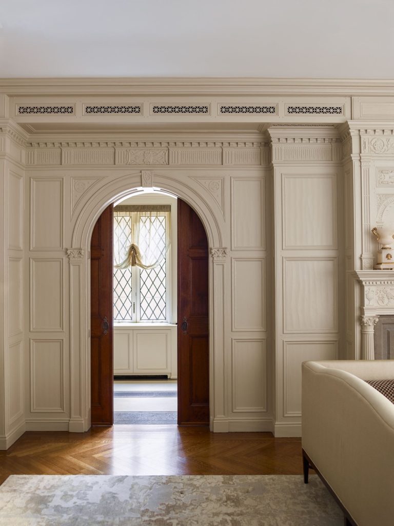 Elegant cream-paneled room with ornate molding and an arched wooden double doorway. Sunlight streams through diamond-pane windows in the hallway beyond. A beige sofa and part of a patterned rug are visible in the foreground.