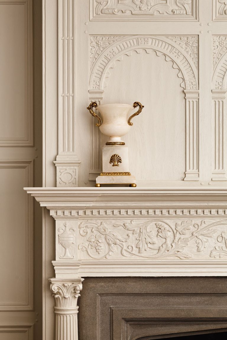 An ornate white marble vase with gold accents sits centered on a carved white mantel. The paneling and mantel feature intricate floral and arch designs, contributing to an elegant, classical interior style.
