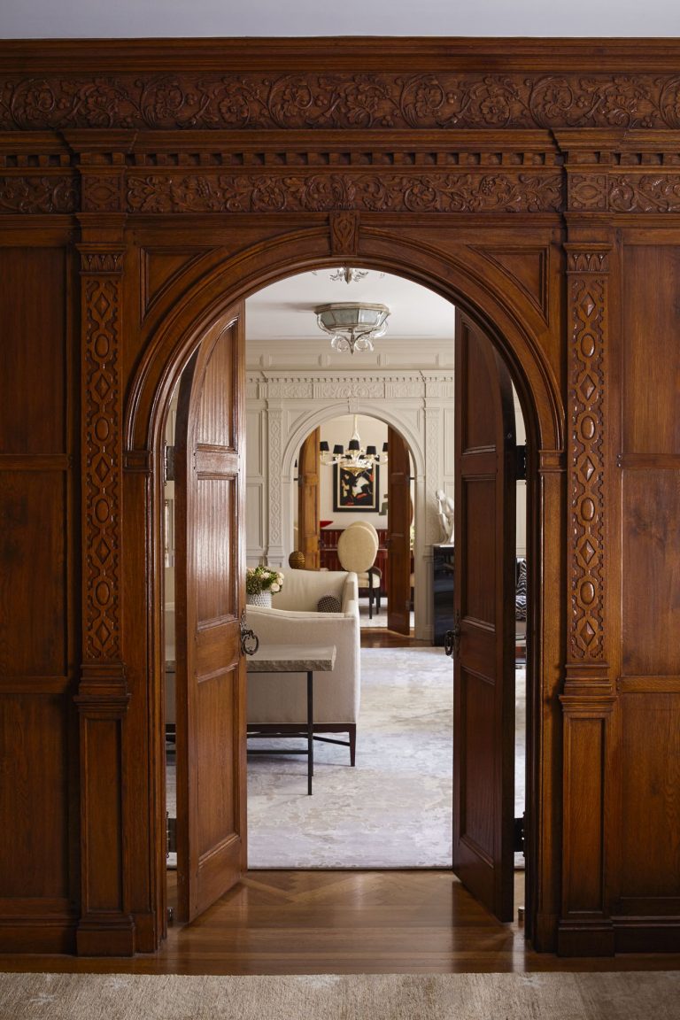 A richly carved wooden archway with open double doors frames a view into an elegant living room, featuring cream-colored walls, a beige sofa, and a painting. Ornate molding and soft lighting add classic, inviting character to the interior.