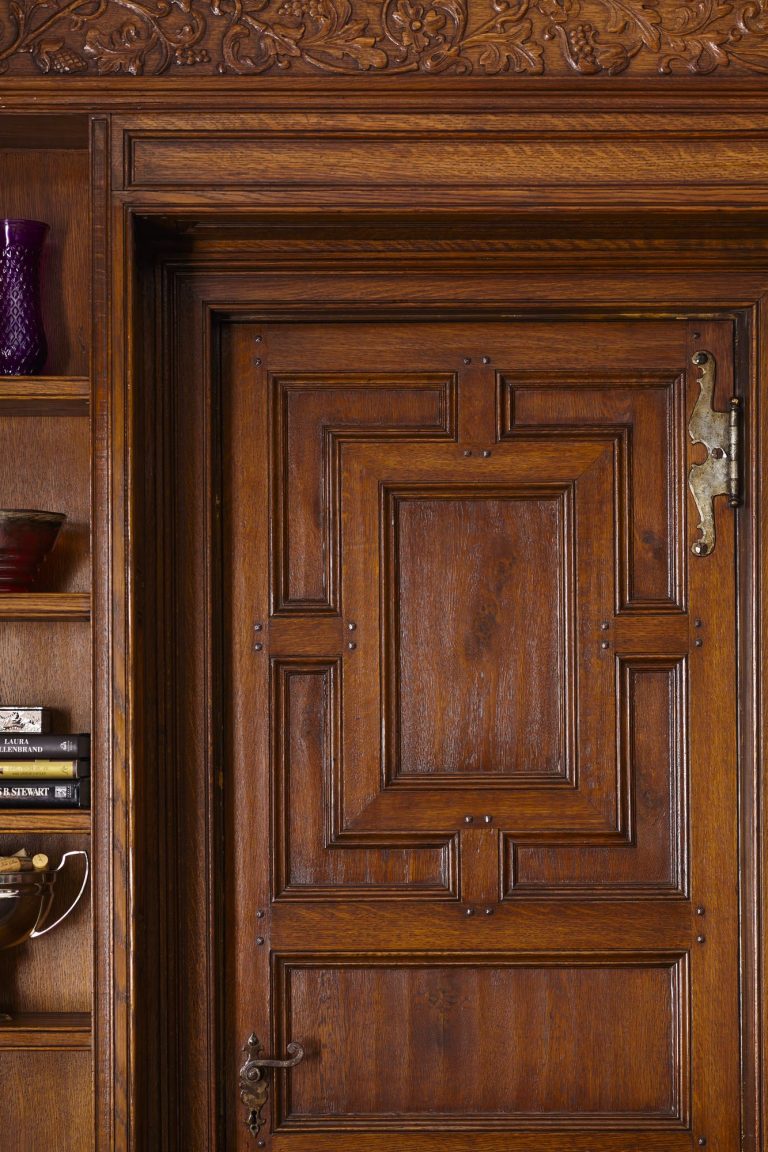 A detailed wooden door with decorative rectangular panels and ornate metal hardware, set in a carved wooden frame. To the left, built-in shelves hold books, bowls, and a purple vase. The wood has a rich, polished finish with intricate grain patterns.