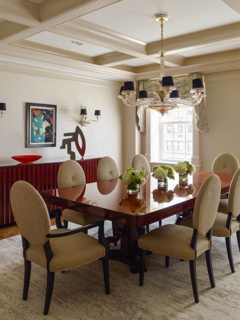 Elegant dining room with a glossy wooden table surrounded by eight beige upholstered chairs. The table is decorated with vases of green hydrangeas. Artwork, red bowl, and modern sculpture sit atop a red sideboard. Large window and chandelier add light.