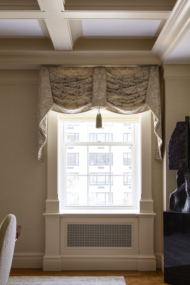 A window with sheer white glass is framed by cream-colored, patterned, draped curtains with a tassel in the center. The room features beige walls, detailed molding, a coffered ceiling, and a decorative radiator cover below the window.
