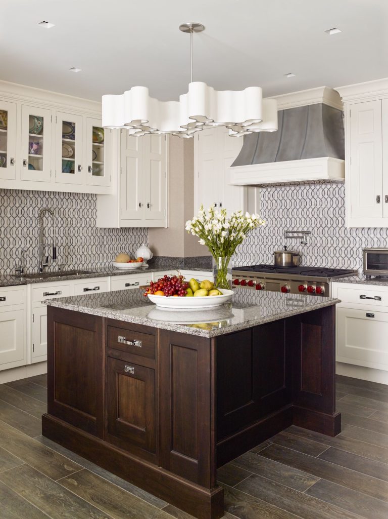 Modern kitchen with white cabinets, patterned tile backsplash, stainless steel range hood, and a dark wood island with a gray stone countertop. A white bowl with apples and grapes and a vase of white flowers sit on the island beneath a sculptural white chandelier.