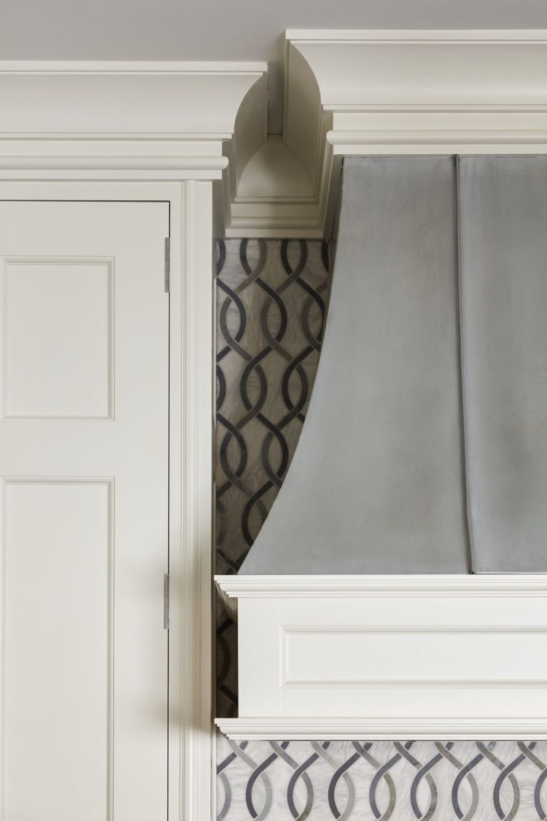 Close-up of a kitchen area showing a white cabinet door with panel detailing on the left, adjacent to a white and gray vent hood with elegant molding. The wall behind has a gray and black wavy, interlocking pattern for a modern touch.