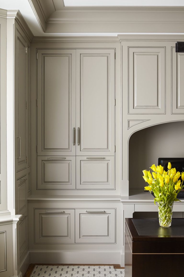 A built-in kitchen cabinet with panel doors in a light beige color, adorned with silver handles, stands next to a countertop holding a vase of bright yellow tulips. A section of dark wood furniture and a patterned floor rug are visible in the foreground.