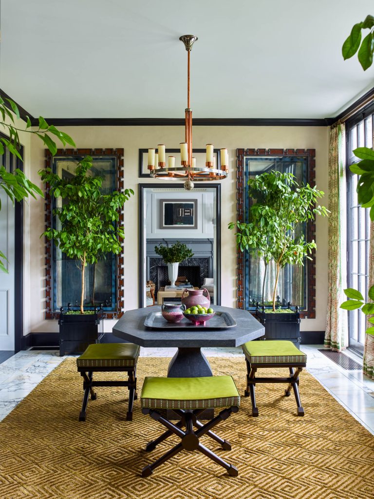 A modern dining room with a hexagonal stone table, four stools with green cushions, a gold geometric rug, large potted plants, tall decorative mirrors, a black fireplace, and a gold chandelier hanging from a white ceiling. Natural light fills the space.