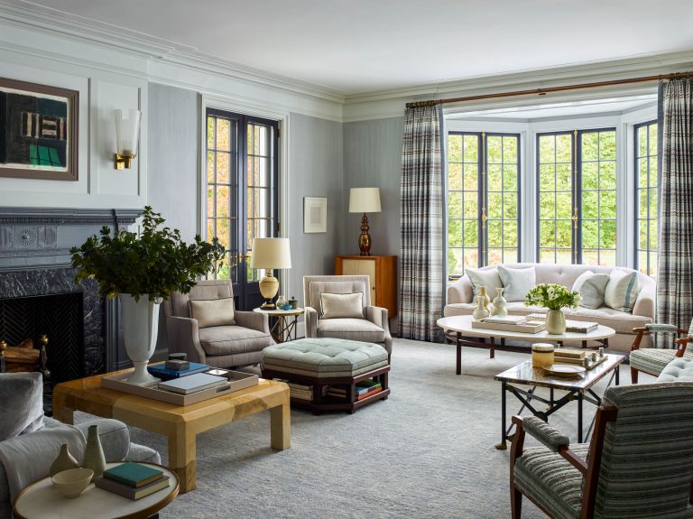Elegant living room with large bay windows, gray walls, and light-filled space. Features neutral sofas and armchairs, a fireplace with art above, patterned drapes, a coffee table with decor, leafy plant, books, and soft, textured furnishings on a pale rug.