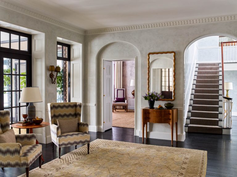 Bright, elegant entryway with patterned armchairs, a wooden table with a mirror and flowers, a beige patterned rug, and grand staircase. Large windows let in natural light; an open doorway reveals another room with a chair and textured wallpaper.