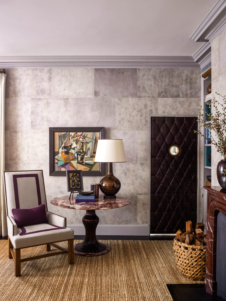 A stylish living room features a marble round table with a glossy lamp, books, and abstract art above it. A modern armchair sits beside the table. The room has textured wallpaper, a quilted brown door, a basket of firewood, and a fireplace on the right.