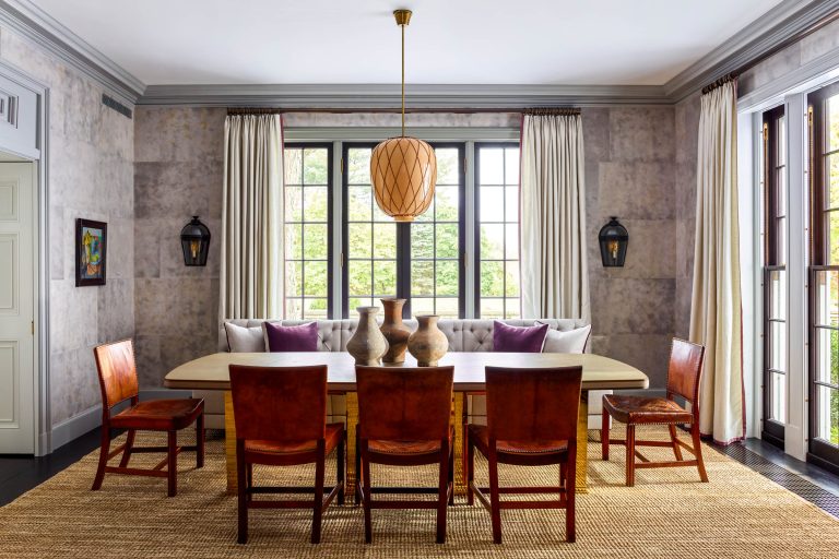 A modern dining room with a large wooden table, six brown chairs, three clay vases as centerpiece, and a cushioned window seat beneath tall windows with cream curtains. Pendant light hangs above, and two wall lanterns are on either side of the window.