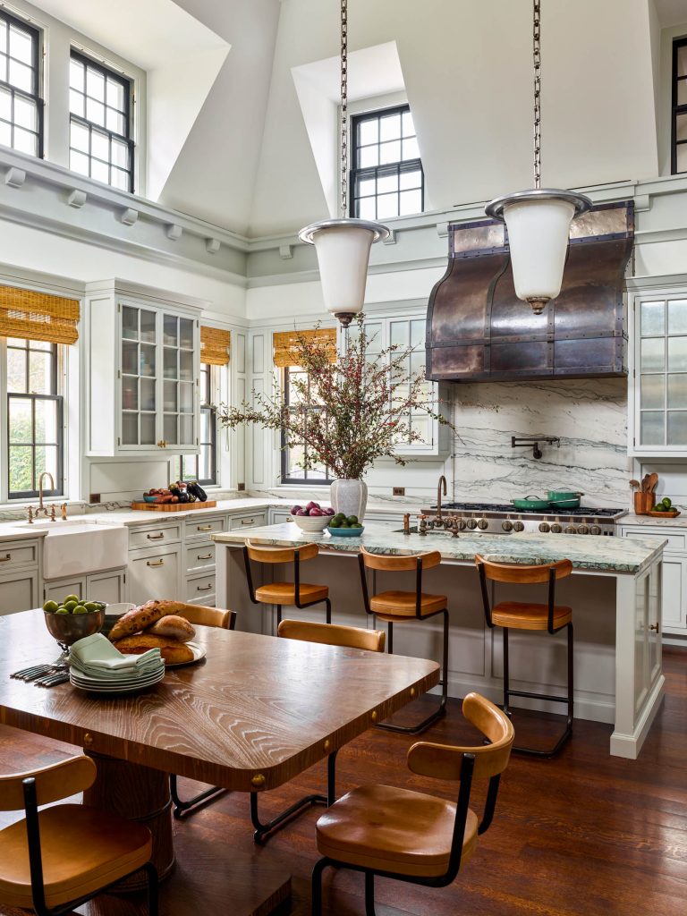 Spacious kitchen with high ceilings, white cabinets, and large windows. Marble island with four wooden bar stools, pendant lights above. Wooden dining table with chairs, fruit bowl, and bread. Vase with branches on the island, copper range hood, hardwood floors.