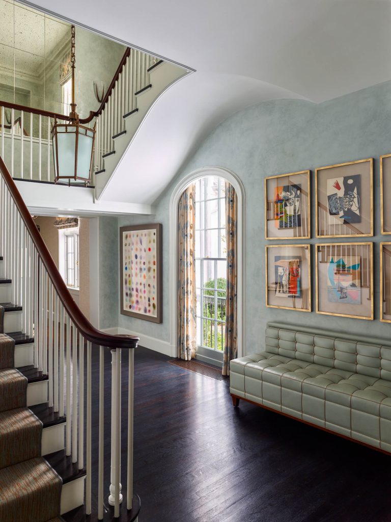 A bright hallway with dark wood floors, a sweeping staircase with white spindles, a tufted green bench, and six framed modern art pieces on a light blue wall. A tall arched window has sheer curtains, and a large lantern-style light fixture hangs above.