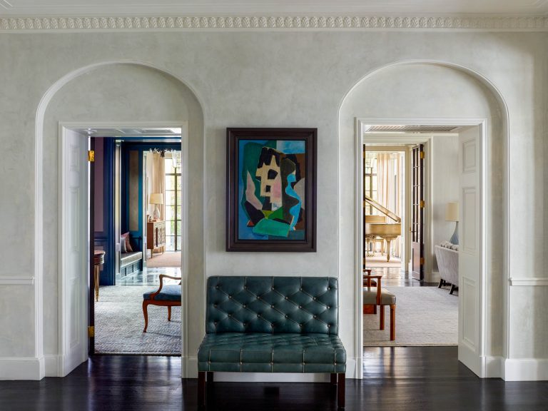A hallway with light gray walls and dark wood floors features a green tufted bench below an abstract painting. Two arched doorways reveal sunlit rooms—one with a grand piano on the right and the other with classic furniture on the left.