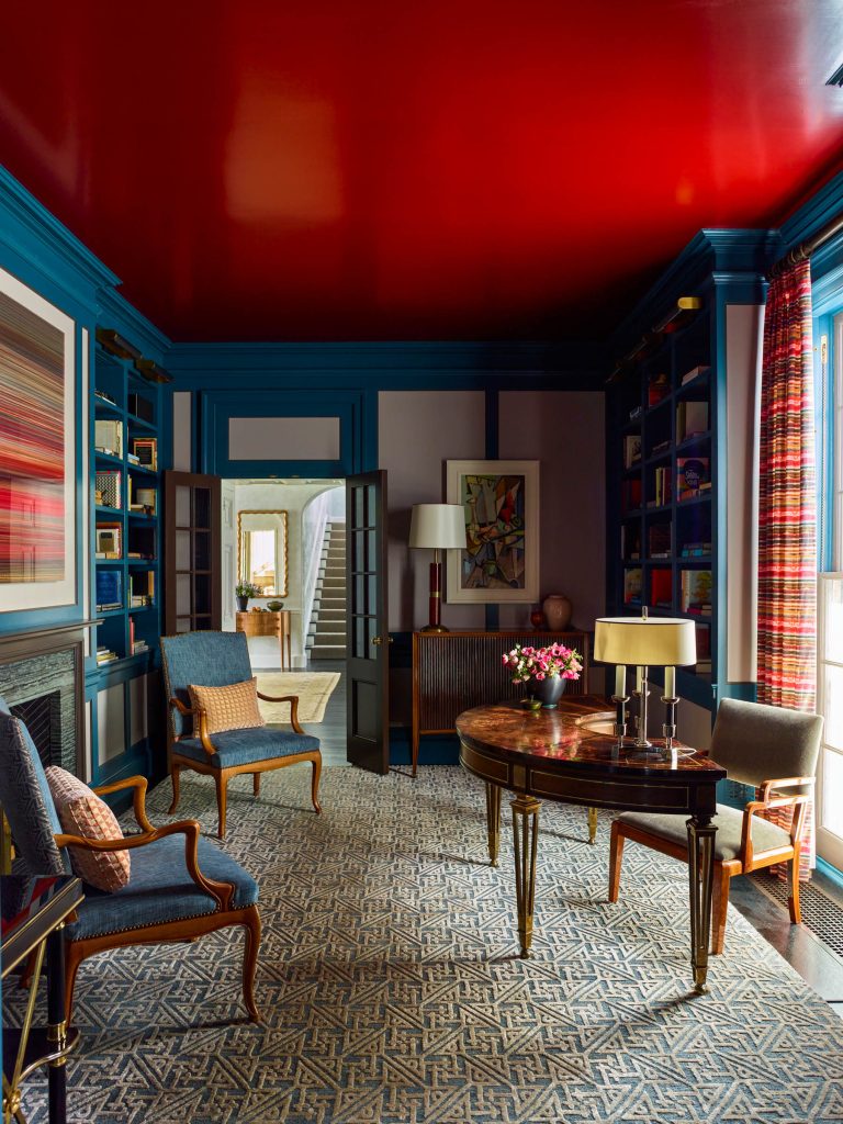 A richly decorated room features a glossy red ceiling, blue paneled walls with built-in bookshelves, elegant armchairs, a round wooden table with two lamps, patterned curtains, a geometric rug, and colorful artwork on the walls.
